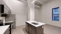 Located on Euclid Avenue in the heart of Cleveland, OH, this one-bedroom, one-bathroom unit offers a thoughtfully designed layout with modern finishes throughout. The open-concept living and kitchen area serves as the centerpiece of the space, featuring exposed ductwork and soaring ceilings that lend an industrial-chic character. The kitchen is outfitted with dark wood cabinetry, stainless steel appliances — including a range, microwave, and refrigerator — and a peninsula island with a white countertop and undermount sink, providing both prep space and a natural gathering point. Light wood-style flooring flows throughout the main living area, complementing the crisp white walls and creating a bright, clean aesthetic. A hallway connects to the private quarters, where the bedroom offers a comfortable footprint with soft carpeting. Adjacent to the bedroom is a generously sized walk-in closet, fully equipped with wire shelving and hanging rods along multiple walls for ample storage. The bathroom features a sleek, all-white standing shower with a sliding glass door, built-in shelving, and polished chrome fixtures, delivering a spa-like feel. Track lighting throughout and a neutral color palette tie the entire unit together with a cohesive, contemporary look.