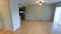 This single-floor residence offers a practical and comfortable layout designed for easy everyday living. The main living area is characterized by clean light hardwood flooring that extends throughout the space, lending a bright and welcoming atmosphere. A ceiling fan with a built-in light fixture is centrally positioned overhead, providing both airflow and ambient lighting. Baseboard heating runs along the wall, ensuring warmth throughout the colder months. The living area flows openly into the kitchen, which is accessed through a wide passthrough opening. The kitchen features dark wood cabinetry that provides generous storage, complemented by black appliances including a range and an overhead microwave. The countertops offer a functional workspace, and the overall layout makes efficient use of the available space. Neutral-toned walls throughout the living area create a versatile backdrop that suits a variety of interior styles. White baseboards provide a crisp contrast against the flooring, adding a finishing touch to the clean aesthetic. The open connection between the living room and kitchen promotes a sense of spaciousness and makes the home well-suited for both relaxed living and casual entertaining. The space is straightforward in design, prioritizing functionality and comfort.