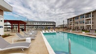 Located at 4710 Old Decatur Rd in Fort Worth, Texas, this outdoor swimming pool area is a well-appointed recreational amenity designed for relaxation and leisure. The centerpiece is a generously sized, crystal-clear pool with a distinctive tiled border, accessible via wide entry steps and handrail access points. A shallow tanning ledge extends into the pool, fitted with in-water lounge chairs — ideal for lounging while staying cool. Surrounding the pool is a spacious, stamped concrete sun deck lined with an abundance of sling-style lounge chairs and occasional side tables, providing ample space for sunbathing. A red-toned pergola structure anchors one side of the deck, offering a shaded gathering area. Additional outdoor dining furniture is available for casual poolside seating. The area is enclosed by a black wrought-iron fence, ensuring privacy and security. Tall pole-mounted lighting fixtures are positioned throughout the deck for evening use. The pool area is flanked on one side by a modern clubhouse building featuring large windows and warm wood-accent paneling, and is surrounded by multi-story residential buildings, creating a cohesive community atmosphere open to the Texas sky.