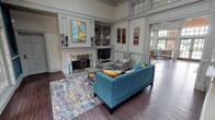 Located at 801–809 W Royal Ln in Irving, Texas, this single-floor community or leasing space offers a refined and welcoming atmosphere defined by thoughtful architectural detailing and high-end finishes throughout. The main lounge area serves as the heart of the space, featuring soaring ceilings with transom windows that draw in abundant natural light. A classic white fireplace with decorative ironwork serves as the focal point, flanked by built-in shelving and cabinetry that provide both display and storage. Rich hardwood flooring runs throughout, complementing the neutral gray paneled walls and crisp millwork. Contemporary furnishings — including a vibrant teal sofa, accent armchairs, and a colorful area rug — add warmth and visual interest to the otherwise classic interior. Adjacent to the lounge, a formal conference and dining area is enclosed in deep navy wainscoting and bathed in natural light from oversized multi-pane windows that frame lush outdoor greenery. A large dark wood table with upholstered chairs makes this room ideal for meetings or gatherings. Floor-to-ceiling French doors with arched transom windows open directly to a furnished outdoor patio, seamlessly blending interior and exterior living. The overall ambiance is polished yet approachable, combining traditional architectural elegance with modern comfort.