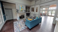Located at 801–809 W Royal Ln in Irving, Texas, this single-floor community or leasing space offers a refined and welcoming atmosphere defined by thoughtful architectural detailing and high-end finishes throughout. The main lounge area serves as the heart of the space, featuring soaring ceilings with transom windows that draw in abundant natural light. A classic white fireplace with decorative ironwork serves as the focal point, flanked by built-in shelving and cabinetry that provide both display and storage. Rich hardwood flooring runs throughout, complementing the neutral gray paneled walls and crisp millwork. Contemporary furnishings — including a vibrant teal sofa, accent armchairs, and a colorful area rug — add warmth and visual interest to the otherwise classic interior. Adjacent to the lounge, a formal conference and dining area is enclosed in deep navy wainscoting and bathed in natural light from oversized multi-pane windows that frame lush outdoor greenery. A large dark wood table with upholstered chairs makes this room ideal for meetings or gatherings. Floor-to-ceiling French doors with arched transom windows open directly to a furnished outdoor patio, seamlessly blending interior and exterior living. The overall ambiance is polished yet approachable, combining traditional architectural elegance with modern comfort.