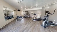 Located in Albuquerque, NM, this fitness center offers a dedicated exercise room designed to support a well-rounded workout routine. The space features durable hardwood-style flooring throughout, providing a clean and functional surface suited for a variety of activities. Track lighting runs along the textured ceiling, delivering bright, focused illumination across the room.

The exercise room is equipped with a comprehensive selection of fitness equipment, including multiple treadmills, an elliptical trainer, a stationary bike, a rowing machine, and a multi-station cable strength machine. A weight bench adds further versatility for strength training. A large wall-mounted mirror spans one side of the room, visually expanding the space and allowing users to monitor their form during workouts. A mounted flat-screen television provides entertainment during cardio sessions.

Natural light filters in through windows on multiple walls, and a full-length sliding glass door opens directly to an outdoor area, offering views of an exterior courtyard and pool. This seamless indoor-outdoor connection enhances the overall atmosphere of the space. The neutral gray and beige wall tones contribute to a clean, focused environment well-suited for fitness activities.