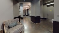 This single-floor lobby space combines modern design with classic touches to create a polished and welcoming entry experience. The expansive reception area is anchored by gleaming polished concrete floors that reflect ambient light, giving the space an open and airy feel. Large glass entry doors flood the interior with natural light, framing street views and enhancing the sense of openness.

At the heart of the lobby sits a sleek front desk finished in deep navy blue, accented by an oversized antique-style skeleton clock and a warm globe wall sconce in brushed brass — details that add character and a timeless quality to the space. A patterned accent chair provides a comfortable waiting area nearby, while dark wainscoting along the lower walls adds depth and contrast to the neutral gray palette throughout.

Moving deeper into the lobby, a corridor leads to a bank of elevators, softly lit by pendant fixtures and flanked by polished concrete columns. An additional service counter area features a metallic credenza topped with glass, displaying vintage black-and-white framed photographs that evoke a sense of history and sophistication. Coffee service amenities are thoughtfully arranged on the counter, suggesting hospitality-oriented use. The overall ambiance is refined, functional, and stylish — well-suited for a professional or hospitality environment.