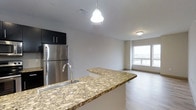 This 2-bedroom, 1-bathroom residence offers a well-designed single-floor layout that balances functionality and modern style. The open-concept kitchen and living area serves as the heart of the home, creating a seamless flow between spaces. The kitchen is highlighted by a sweeping granite countertop peninsula with warm earth-tone veining, providing ample prep space and a natural gathering point. Dark cabinetry overhead contrasts with lighter elements throughout, while stainless steel appliances, including a full-size refrigerator, add a sleek, contemporary feel. A pendant light fixture hangs above the peninsula, contributing both task lighting and visual interest. The living area extends naturally from the kitchen, bathed in natural light from a large window that brightens the space and offers outward views. Light-toned hardwood-style flooring runs throughout, lending warmth and visual continuity across the open floor plan. The neutral wall palette keeps the space feeling bright and versatile. Overall, the residence presents a clean, modern aesthetic with quality finishes that make it both practical and inviting.