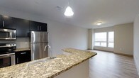 This 2-bedroom, 1-bathroom residence offers a well-designed single-floor layout that balances functionality and modern style. The open-concept kitchen and living area serves as the heart of the home, creating a seamless flow between spaces. The kitchen is highlighted by a sweeping granite countertop peninsula with warm earth-tone veining, providing ample prep space and a natural gathering point. Dark cabinetry overhead contrasts with lighter elements throughout, while stainless steel appliances, including a full-size refrigerator, add a sleek, contemporary feel. A pendant light fixture hangs above the peninsula, contributing both task lighting and visual interest. The living area extends naturally from the kitchen, bathed in natural light from a large window that brightens the space and offers outward views. Light-toned hardwood-style flooring runs throughout, lending warmth and visual continuity across the open floor plan. The neutral wall palette keeps the space feeling bright and versatile. Overall, the residence presents a clean, modern aesthetic with quality finishes that make it both practical and inviting.
