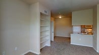 This single-floor space offers a straightforward, functional layout highlighted by several practical design elements. The main living area features wall-to-wall carpeting in a neutral tone, contributing to a warm and understated ambiance throughout. A standout feature is the built-in bookshelf unit recessed into the wall, offering multiple open shelves ideal for storage or display. The walls are finished in a light beige tone, lending a clean and cohesive look to the interior. Overhead ventilation registers are visible, indicating a central HVAC system for year-round climate control. A flush-mount ceiling light fixture provides overhead illumination in the adjoining room, which is accessible through a dark-toned interior door. To the right, a partial view of white cabinetry suggests a kitchen or utility area nearby, adding to the overall functionality of the space. White baseboards run along the perimeter, providing a crisp contrast to the carpeted floors. The open sightlines between areas enhance the sense of connectivity within the layout, making the space feel cohesive and easy to navigate.