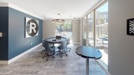 Located at 10001 E Dry Creek Rd in Englewood, Colorado, this single-floor amenity space is thoughtfully designed to serve both productivity and recreation. The layout flows seamlessly across distinct zones, each with its own character and purpose.

The collaborative work area is bright and inviting, featuring floor-to-ceiling windows that frame views of an outdoor pool and surrounding grounds, flooding the space with natural light. A round conference table surrounded by rolling upholstered chairs sits beneath a sculptural branch-style chandelier, while a bold blue accent wall with a graphic "R" logo adds a vibrant, branded touch. A bistro-height table near the windows provides an additional spot for casual work or conversation.

A well-appointed kitchenette serves the space with clean, white cabinetry, light wood countertops, an integrated sink, a stainless steel microwave, and a mini-refrigerator. The wall above is adorned with a striking arrangement of natural wood cross-section art pieces, lending an organic, artistic element to the contemporary design. A glass-panel sliding door connects this area to the rest of the space.

The game room is anchored by a full-size ping-pong table and a shuffleboard table, set against a dramatic dark navy accent wall featuring a large architectural photomural. A wall-mounted television completes the entertainment setup. Durable wood-look flooring runs throughout, tying the entire space together with a modern, cohesive aesthetic.