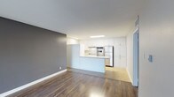This well-appointed single-floor space showcases a clean, modern design with an open-concept layout that seamlessly connects the living area to the kitchen. The kitchen is a standout feature, highlighted by crisp white cabinetry that extends from counter height to the ceiling, offering generous storage. Stainless steel appliances — including a built-in oven, stovetop, and refrigerator — lend a polished, contemporary feel. A peninsula counter serves as a natural divider between the kitchen and the open living area, making it ideal for casual dining or meal prep. Rich, dark hardwood flooring flows throughout the space, providing a warm contrast to the bright white cabinetry and neutral grey accent wall. The living area is open and versatile, benefiting from a clean, minimalist aesthetic with white walls and smooth ceilings featuring a skylight panel that draws in natural light. The overall ambiance is modern and functional, with a neutral palette that offers a flexible canvas for a variety of interior styles. The thoughtful layout and quality finishes make this a practical and visually appealing space.