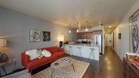 This single-floor residence is thoughtfully designed with a contemporary aesthetic that balances functionality and style. The open-concept layout seamlessly connects the living area to the kitchen, creating a cohesive and sociable space. The living room features dark hardwood-style flooring, large windows that invite natural light, and direct access to an outdoor area, offering views of the surrounding building exterior. The neutral wall tones provide a versatile backdrop for varied décor styles.

The kitchen is well-appointed with warm walnut-toned cabinetry, stainless steel appliances including a French door refrigerator, electric range, and built-in microwave. A peninsula with bar seating and a light-colored countertop serves as both a practical workspace and an informal dining spot. Track lighting and pendant fixtures illuminate the area effectively.

The bedroom features plush carpeting, a ceiling fan, and a sliding-door closet, creating a comfortable and functional retreat. The bathroom is finished with dark hardwood-style flooring, a walnut-toned vanity with a grey countertop, and a shower-tub combination, maintaining the modern design language found throughout the residence.