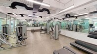 Located at 100 Seventh St in downtown Pittsburgh, this fitness center offers a comprehensive workout environment across a single, open floor. The space is thoughtfully designed with distinct zones for cardio and strength training, providing ample room to move between equipment. The cardio area features a row of treadmills, elliptical machines, stationary bikes, and a rowing machine, with a mounted flat-screen TV for entertainment during workouts. The strength training zone is equally well-appointed, housing a full dumbbell rack with benches, a variety of cable and resistance machines, and a functional training station with resistance bands and medicine balls. Floor-to-ceiling glass partitions divide sections of the space while maintaining an open, airy feel, and wall-to-wall mirrors amplify the sense of spaciousness throughout. Large-format tile flooring and rubberized mat areas in the free weights section provide both durability and safety. The exposed ceiling with industrial-style ductwork and bright fluorescent lighting lend the space a modern, no-frills aesthetic. The overall design is clean, functional, and well-lit, making it a practical and inviting facility for a full-body workout routine.