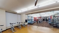 This single-floor fitness center is thoughtfully designed to accommodate a range of workout activities within a clean, modern environment. The space is divided into two distinct zones, separated by a full-height glass partition that maintains an open, connected feel while defining each area's purpose.

The cardio zone houses a row of treadmills positioned along one wall, allowing users ample space to exercise comfortably. Warm wood-tone flooring runs throughout this area, contributing to a welcoming atmosphere. Bright fluorescent lighting is installed across the ceiling in linear arrangements, ensuring the entire space is evenly and thoroughly illuminated.

Adjacent to the cardio area, a dedicated cycling zone features several stationary bikes arranged in a row, suitable for individual or group cycling sessions. A projection screen is mounted on the wall nearby, adding a multimedia element to workout sessions.

The strength training section, visible through the glass partition, is equipped with cable machines, weight racks, and various resistance equipment. Red accent walls in this zone add a energetic visual contrast to the otherwise neutral palette. The overall design emphasizes functionality and clarity, with an efficient layout that maximizes usable floor space throughout the facility.