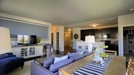 This single-floor residence showcases an open-concept layout that connects the living, dining, and kitchen areas into one cohesive and functional space. The living area serves as the focal point, anchored by a plush blue sectional sofa accented with nail-head trim and layered with coordinating throw pillows. A striped blue-and-white area rug grounds the seating arrangement, while a rustic wood coffee table adds warmth and contrast. An accent wall featuring a painted rectangular molding detail frames the mounted flat-screen television, adding a touch of architectural character. Built-in shelving nearby provides both storage and display space. The open-concept design flows naturally into the kitchen, which features dark espresso cabinetry, light countertops, and a peninsula with bar-height seating — ideal for casual dining or entertaining. Pendant-style lighting illuminates the kitchen area, complementing the recessed lighting throughout. Dark hardwood flooring runs consistently across the space, tying the areas together cohesively. The neutral wall tones, accented by cool blues and deep wood finishes, create a balanced, inviting ambiance throughout the home.