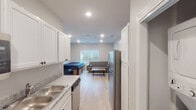 Located at 1607 Blackjack Rd in Starkville, Mississippi, this single-floor unit offers a clean, modern design with a functional open-concept layout. Light hardwood-style flooring runs throughout, creating a cohesive and bright aesthetic complemented by recessed lighting and neutral gray-and-white walls.

The kitchen is well-appointed with crisp white shaker cabinetry, granite-look countertops, a stainless steel refrigerator, dishwasher, and range. Ample upper and lower cabinet storage lines both sides of the galley-style kitchen, which flows seamlessly toward the main living area.

The combined living and sleeping space is generously sized, accommodating a sofa, coffee table, wall-mounted television with a media stand, and a bed with built-in storage drawers beneath. A window fitted with blinds allows natural light to brighten the room.

A walk-in closet with built-in shelving and rod provides organized storage. The bathroom includes white cabinetry, a sink vanity, and a full tub-shower combination with brushed nickel fixtures. The overall layout is efficient and well-designed, making thoughtful use of the available space throughout.