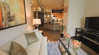 This single-floor space showcases an open-concept layout that connects the living area and kitchen in a cohesive, loft-inspired design. The polished concrete floors reflect ambient light throughout, lending a sleek, industrial character to the interior. Exposed ductwork along the ceiling reinforces the urban aesthetic, while track lighting and elegant pendant lights provide layered illumination across the space.

The living area is anchored by a plush cream sofa accented with sage green pillows, a glass-and-metal coffee table, and a soft area rug, creating a comfortable yet contemporary seating arrangement. A warm table lamp adds a cozy glow alongside the track lighting above.

The kitchen is thoughtfully designed with warm honey-toned wood cabinetry, a decorative mosaic tile backsplash, and stainless steel appliances. A central island with a dark countertop serves as a functional hub, ideal for meal preparation or casual dining. Pendant lights suspended above the island add a refined visual element.

Overall, the space balances industrial and warm design elements, creating a stylish and functional environment with a modern urban ambiance.