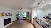 This well-appointed single-floor residence offers a thoughtfully designed open layout that seamlessly connects the living, dining, and kitchen areas. Rich hardwood flooring flows throughout the main living spaces, lending warmth and continuity to the interior.

The living area is a standout, anchored by a classic white-mantled fireplace that serves as a natural focal point. A ceiling fan overhead and large windows allow ample natural light to filter in, offering views of lush greenery outside. A glass door provides access to an outdoor area, further enhancing the connection between indoor and outdoor living.

The kitchen is efficiently organized in a galley-style configuration, featuring white shaker cabinetry, dark granite countertops, and a full suite of stainless steel appliances including a refrigerator, oven, microwave, and gas range. Track lighting and recessed fixtures illuminate the space effectively. A double-basin sink with a modern faucet is positioned along the counter, offering a practical workspace.

Adjacent to the living area, a dedicated dining space provides room for casual meals, while a quiet corner with a wooden desk creates a comfortable area for work or study. The overall ambiance of the space is clean, modern, and inviting, with a neutral color palette of white walls and warm wood tones that creates a calm, cohesive aesthetic throughout.