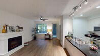 This well-appointed single-floor residence offers a thoughtfully designed open layout that seamlessly connects the living, dining, and kitchen areas. Rich hardwood flooring flows throughout the main living spaces, lending warmth and continuity to the interior.

The living area is a standout, anchored by a classic white-mantled fireplace that serves as a natural focal point. A ceiling fan overhead and large windows allow ample natural light to filter in, offering views of lush greenery outside. A glass door provides access to an outdoor area, further enhancing the connection between indoor and outdoor living.

The kitchen is efficiently organized in a galley-style configuration, featuring white shaker cabinetry, dark granite countertops, and a full suite of stainless steel appliances including a refrigerator, oven, microwave, and gas range. Track lighting and recessed fixtures illuminate the space effectively. A double-basin sink with a modern faucet is positioned along the counter, offering a practical workspace.

Adjacent to the living area, a dedicated dining space provides room for casual meals, while a quiet corner with a wooden desk creates a comfortable area for work or study. The overall ambiance of the space is clean, modern, and inviting, with a neutral color palette of white walls and warm wood tones that creates a calm, cohesive aesthetic throughout.