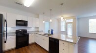 This single-floor residence offers a well-designed open-concept layout that maximizes both functionality and natural light. The kitchen serves as the heart of the home, showcasing crisp white shaker-style cabinetry paired with granite countertops that extend across a generous peninsula with an undermount sink. Black appliances, including a range, microwave, and dishwasher, provide a striking contrast against the clean white cabinetry. A trio of pendant lights hangs above the peninsula, complementing the flush ceiling fixture to create layered, practical lighting throughout the kitchen. Rich hardwood flooring runs throughout, adding warmth and continuity to the space. The kitchen opens directly into the living area, which benefits from abundant natural light streaming through large windows dressed with blinds. A chandelier anchors the living space, adding an elegant touch. The seamless flow between the kitchen and living areas makes the layout ideal for everyday living and entertaining. Overall, the space presents a clean, modern aesthetic characterized by bright interiors, quality finishes, and a cohesive design that balances style with practicality.