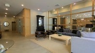 This single-floor space showcases a thoughtfully designed lounge or reception area that blends contemporary and classic design elements. The room is anchored by a large grid-framed mirror wall constructed with warm wood framing, which adds depth and amplifies the sense of space while reflecting the interior's carefully chosen furnishings. Recessed lighting throughout the ceiling provides consistent, clean illumination across the space.

The seating area features a mix of rich brown leather armchairs with chrome bases and a plush gray tufted sofa, arranged around a rectangular white coffee table and grounded by a neutral woven area rug. A striking dark wood display cabinet with glass panels serves as a dramatic focal point against the light-toned walls. Adjacent to the seating area, a tall wood-paneled column adds a natural, textural contrast to the otherwise smooth wall surfaces.

The overall ambiance is polished and welcoming, balancing warm wood tones with neutral upholstery and sleek metallic accents. The open layout and strategic use of mirrors contribute to an airy, expansive feel suited for a professional or hospitality-oriented environment.