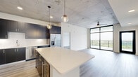 This contemporary 2-bedroom, 2-bathroom residence showcases an industrial-modern aesthetic defined by dramatic exposed concrete ceilings, light wood-look flooring, and bold black-framed architectural details throughout. The open-concept kitchen and living area forms the heart of the home, where a large white quartz island anchors the space beneath globe pendant lights. The kitchen is outfitted with dark navy cabinetry, stainless steel appliances including a French door refrigerator, gas range, microwave, and dishwasher, all complemented by crisp white countertops. The expansive living area is bathed in natural light through floor-to-ceiling windows that open to a private balcony, offering open outdoor views. A ceiling fan adds both style and function to the living space. An exposed concrete accent wall adds an artistic, industrial touch. The primary bedroom is generously sized, featuring direct balcony access via sliding glass doors, a ceiling fan, and abundant natural light. The primary bathroom offers a dual-sink vanity with bold blue cabinetry, white quartz counters, and a walk-in glass shower. Each bedroom is served by a dedicated closet with wire shelving systems. The secondary bedroom also features exposed concrete ceiling details. Three hallways connect the layout efficiently, and the overall palette of white walls, light flooring, and contrasting dark fixtures creates a refined, cohesive living environment.