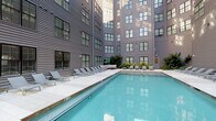This amenity space is comprised of two distinct areas: an outdoor pool and a comprehensive fitness center. The outdoor pool is set within a courtyard framed by a multi-story building with gray horizontal siding and large black-trimmed windows. The pool features clear, blue-tinted water and is surrounded by a spacious concrete deck furnished with multiple lounge chairs, providing ample room for relaxation. Tropical plantings add a touch of greenery to the setting.

The fitness center is a bright, well-lit space finished with neutral gray walls, recessed lighting, and a ceiling fan that enhances air circulation. Large windows and glass sliding doors allow abundant natural light to filter in and offer views of the outdoor pool area. Floor-to-ceiling mirrors line the walls, visually expanding the space. The facility is thoroughly equipped with treadmills, ellipticals, and stationary bikes for cardio training, as well as multi-station cable machines, adjustable benches, a full dumbbell rack, and a medicine ball station. Wooden open shelving provides storage for towels and accessories, contributing to an organized and polished aesthetic throughout.