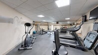 Located at 13401 Morgan Ave S in Burnsville, Minnesota, this fitness center offers a well-equipped, single-floor facility designed to support a well-rounded workout routine. The space is divided into two dedicated exercise rooms, providing a functional layout that separates cardio and strength training areas.

The cardio area is outfitted with multiple treadmills, each equipped with individual monitors for entertainment during workouts, as well as elliptical machines and a stationary bike. Wall-mounted flat-screen televisions add to the amenities, making extended cardio sessions more engaging. The flooring throughout is durable rubber fitness matting, ideal for both safety and equipment support.

The strength training area features a multi-station cable machine, a weight bench with a barbell rack, a dumbbell rack stocked with free weights, and a stability ball, offering a comprehensive selection of resistance training options. A floor-to-ceiling mirror lines one wall, enhancing the sense of space and allowing users to monitor their form. A wall-mounted air conditioning unit ensures a comfortable environment year-round.

The facility features a drop ceiling with recessed panel lighting that provides bright, even illumination throughout. The neutral, warm-toned walls create a clean and focused atmosphere. An additional room off the main exercise areas provides supplemental space. Overall, the fitness center is a compact yet thoroughly equipped facility suited for a diverse range of fitness needs.