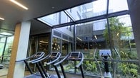 This contemporary fitness center is designed to seamlessly blend indoor exercise with the natural environment. The space is defined by expansive floor-to-ceiling glass walls and a generous skylight overhead, flooding the interior with natural light and creating an open, airy atmosphere. A row of modern treadmills, each equipped with personal digital displays, is positioned directly facing the glass facade, offering exercisers an engaging view of a lush, landscaped courtyard garden below. The greenery of the garden — featuring a variety of tropical-style plants — provides a calming visual contrast to the sleek, urban architectural surroundings visible beyond the glass. The structural framework of the space incorporates dark steel accents, complementing the building's modern aesthetic. Beyond the fitness floor, additional areas of the facility are visible through interior glass partitions, suggesting a well-appointed, multi-functional wellness space. The overall ambiance strikes a balance between high-performance functionality and a refined, design-forward environment, making it a standout amenity within what appears to be a larger urban residential or mixed-use development.