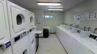 This single-floor communal laundry facility is designed for efficiency and convenience, offering a well-organized layout with all the essentials for a complete laundry experience. The room is lined with a row of top-loading washing machines along one wall, each equipped with individual controls and dispensers, providing ample capacity for multiple users simultaneously. On the opposite side, stacked washer and dryer units maximize the use of vertical space, further increasing the facility's overall capacity. A utility sink is conveniently positioned along the back wall, accompanied by a wall-mounted dispenser, offering a practical solution for hand-washing or pre-treating garments. A small folding table is centrally placed, providing a dedicated surface for sorting and folding laundry. The space is illuminated by bright overhead fluorescent lighting, ensuring excellent visibility throughout. Light gray walls and a neutral-toned floor contribute to the clean, utilitarian aesthetic of the room. A small high window along the back wall allows a touch of natural light to filter in. The facility also includes a waste bin for added convenience, keeping the space tidy and well-maintained.
