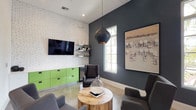 This contemporary single-floor space offers a thoughtfully designed lounge area that seamlessly blends industrial and modern aesthetics. The interior is anchored by polished concrete floors that reflect the abundant natural light streaming in through large, multi-pane windows overlooking lush greenery outside. Soaring ceilings enhance the sense of openness, while a dramatic matte black pendant light serves as a striking focal point above a natural wood stump coffee table at the heart of the seating area. The lounge features a curated arrangement of charcoal gray wingback chairs positioned around the central table, encouraging conversation and relaxation. A whitewashed exposed brick wall provides textural contrast against the deep slate-gray accent wall, where framed artwork adds a gallery-like touch. Built-in lime-green cabinetry beneath a wall-mounted flat-screen TV injects a vibrant pop of color, complemented by floating industrial shelves displaying decorative accessories. A compact stainless steel refrigerator is integrated into the cabinetry, adding functionality to the space. A connecting hallway, illuminated by a wall sconce, leads to an adjacent room visible in the background, suggesting a multi-room layout designed for both comfort and utility. The overall ambiance is modern, energetic, and welcoming.