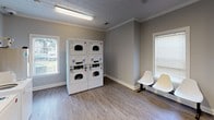 Located at 5577 Houston Rd in Macon, Georgia, this single-floor community laundry facility is designed to provide a clean, functional, and comfortable experience for its users. The space features durable wood-look vinyl flooring, neutral gray walls, and crown molding throughout, lending a tidy and well-kept atmosphere.

The main laundry area houses a row of multiple top-load washing machines, each numbered and equipped with CSC ServiceWorks payment systems, enabling convenient cashless payment options. Instructional and service signage is prominently displayed along the walls for easy reference. A set of stacked dryer units provides ample drying capacity alongside the washers.

Natural light filters into the space through several windows fitted with blinds, complemented by overhead fluorescent lighting that ensures the area remains bright and functional at all hours. A folding table along one wall offers a practical surface for sorting and folding laundry.

The facility includes a dedicated seating area with white chairs, allowing users to wait comfortably. A clearly marked entrance door with a window panel provides easy access, while a waste bin and "No Smoking" signage reflect a commitment to cleanliness and user consideration. The overall layout is open and efficiently organized, making this laundry facility a practical and welcoming amenity.