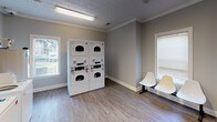 Located at 5577 Houston Rd in Macon, Georgia, this single-floor community laundry facility is designed to provide a clean, functional, and comfortable experience for its users. The space features durable wood-look vinyl flooring, neutral gray walls, and crown molding throughout, lending a tidy and well-kept atmosphere.

The main laundry area houses a row of multiple top-load washing machines, each numbered and equipped with CSC ServiceWorks payment systems, enabling convenient cashless payment options. Instructional and service signage is prominently displayed along the walls for easy reference. A set of stacked dryer units provides ample drying capacity alongside the washers.

Natural light filters into the space through several windows fitted with blinds, complemented by overhead fluorescent lighting that ensures the area remains bright and functional at all hours. A folding table along one wall offers a practical surface for sorting and folding laundry.

The facility includes a dedicated seating area with white chairs, allowing users to wait comfortably. A clearly marked entrance door with a window panel provides easy access, while a waste bin and "No Smoking" signage reflect a commitment to cleanliness and user consideration. The overall layout is open and efficiently organized, making this laundry facility a practical and welcoming amenity.