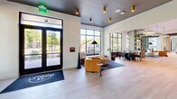The Brinkley Lobby at 3010 Edgeview Ln is a thoughtfully designed 2,540 square foot single-floor communal space that blends modern aesthetics with functional versatility. The main entry welcomes visitors with light hardwood-style flooring, soaring ceilings, and floor-to-ceiling black-framed windows that flood the interior with natural light and offer views of surrounding greenery. Exposed concrete columns and decorative wood lattice screens add architectural character throughout. The open lounge area features stylish seating arrangements, including a mustard-yellow sofa, built-in booth seating with warm wood paneling, and open shelving units, creating a welcoming social environment. A dedicated dining area with pendant lighting provides a space for more structured gatherings. Multiple private meeting rooms are available, each equipped with conference tables, comfortable seating, wall-mounted displays, and globe or industrial-style pendant lighting. One meeting room features a large window overlooking the exterior landscape, while another includes built-in cabinetry. A smaller breakout room with a round table and leather chairs offers an intimate setting for focused conversations. The overall ambiance is contemporary and professional, combining warm tones, curated furnishings, and abundant natural light.