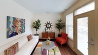 This single-floor residence showcases a cohesive mid-century modern design throughout, combining warm wood tones, bold artwork, and an inviting color palette. The living area serves as the heart of the space, featuring rich hardwood-style flooring, a clean white sofa accented with colorful throw pillows, and an eye-catching hexagonal coffee table. Lush fiddle-leaf fig trees and vibrant wall art lend the room a lively, curated character, while large windows invite ample natural light.

The kitchen is well-appointed with warm maple cabinetry, dark granite countertops, a classic subway tile backsplash in a soft blue tone, and a full suite of black appliances including a range, microwave, refrigerator, and dishwasher. Track lighting overhead ensures the workspace is bright and functional.

The bedroom is a tranquil retreat, offering hardwood-style flooring, sheer-curtained windows that allow in soft natural light, a ceiling fan, and thoughtfully selected furnishings. Colorful artwork adds personality without overwhelming the calm atmosphere.

The bathroom is clean and practical, featuring a single-sink vanity with wood cabinetry, a tub-shower combination, and tile flooring. Near the entry, a convenient coat closet and a compact dining nook with teal accent seating complete this well-designed and characterful space.