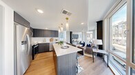 This thoughtfully designed single-floor residence showcases a contemporary aesthetic throughout, with warm hardwood flooring, clean white walls, and black hardware accents that create a cohesive, modern feel. The open-concept kitchen and living area serves as the heart of the home, anchored by a generous center island with a crisp white quartz countertop and bar seating. The kitchen features sleek dark cabinetry, stainless steel appliances, a gold-toned faucet, and a white tile backsplash. Elegant pendant lighting overhead adds a refined touch. The living area flows naturally from the kitchen and is bathed in natural light from floor-to-ceiling windows that frame urban cityscape views and connect to an exterior balcony. The primary bedroom offers a calm retreat with hardwood flooring, recessed lighting, and access to abundant natural light. The en-suite bathroom is finished with floor-to-ceiling marble-style tile, a glass-enclosed walk-in shower with dark hexagonal floor tile, and matte black fixtures. A second bathroom features a soaking tub with marble-style surround, hexagonal floor tile, a floating vanity, and matching black hardware. A bright, light-filled hallway with large windows connects the rooms, completing this well-appointed urban residence.