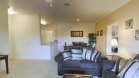 This single-floor residence offers a thoughtfully designed living space that balances comfort and style. The living room serves as the heart of the home, featuring plush, dark upholstered sofas arranged around a glass-top coffee table, creating an inviting area for relaxation and entertaining. A colorful area rug anchors the seating arrangement, adding warmth and visual interest to the space. Crown molding details along the ceiling lend a polished, refined touch to the room, while a ceiling fan ensures year-round comfort. Decorative wall art adds personality and character to the neutral-toned walls. A standout feature of the living area is the large sliding glass door, which floods the room with natural light and opens directly onto a private balcony. The balcony is outfitted with outdoor seating, offering a pleasant spot to enjoy the surrounding views. Sheer curtains and valances frame the glass door, softening the transition between indoor and outdoor living. The overall ambiance is warm and residential, with a layout that makes efficient use of the available space while maintaining an open, airy feel throughout.
