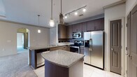 This single-floor residence offers a thoughtfully designed layout that balances modern finishes with comfortable living. The kitchen serves as a central highlight, showcasing dark espresso cabinetry extending to the ceiling, granite countertops, and a full suite of stainless steel appliances including a range, built-in microwave, dishwasher, and french door refrigerator. A curved kitchen island provides additional prep space and casual seating, while pendant lights and track lighting illuminate the room. The kitchen opens to the living area through a peninsula counter, creating a connected, open feel. The living space features soft carpeting, crown molding, and an arched passageway that adds architectural character. A separate room with a tray ceiling, built-in shelving, ceiling fan, and a glass door leading to a balcony offers versatile use as a living or dining area with access to natural light. The bedrooms are carpeted and feature large windows with blinds, ceiling fans, and access to walk-in closets. The bathroom is finished with granite-topped dark wood vanities, large-format tile flooring, and a subway-tiled soaking tub and shower combination.