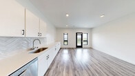Located at 155 Jackson St in Oshkosh, Wisconsin, this thoughtfully designed studio unit offers a clean, contemporary aesthetic throughout its efficiently arranged single-floor layout. The open-concept main area seamlessly integrates the kitchen, dining, and living spaces, creating a bright and cohesive environment ideal for modern living. The kitchen is outfitted with sleek white flat-front cabinetry accented by matte black hardware, light quartz countertops, a stainless steel sink, a herringbone-patterned tile backsplash, and stainless steel appliances including a dishwasher and built-in microwave. Recessed lighting and black-framed windows and entry door add a sharp, contemporary contrast against the crisp white walls. Wood-look luxury vinyl plank flooring runs consistently throughout the entire unit, contributing to a warm yet modern feel. The bedroom area is defined by a stylish barn-door closet with built-in shelving for organized storage. The bathroom maintains the same clean design language, featuring a white vanity with matching cabinetry and wood-look flooring. A private balcony extends the living space outdoors, accessible through the glass-paneled entry door, offering an additional area to enjoy the surroundings.