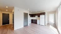 This single-floor unit presents a clean, contemporary design with light hardwood-style flooring flowing consistently throughout the space. The open-concept layout connects the kitchen and living area seamlessly, creating a functional and airy environment.

The kitchen is equipped with dark espresso cabinetry extending to the ceiling, offering generous storage. White appliances — including a refrigerator, range, microwave, and dishwasher — provide a crisp contrast against the dark cabinetry. Light countertops and a stainless steel sink complete the practical workspace. A louvered interior door adds a subtle design detail near the kitchen transition.

The living area is notably bright, benefiting from large floor-to-ceiling windows that frame urban rooftop views and flood the space with natural light. Recessed lighting supplements the natural illumination throughout.

The bathroom features warm-toned hardwood-style flooring, a full soaking tub with shower and curtain rod, a toilet, and a wide vanity with dark cabinetry matching the kitchen, topped with a light countertop and single sink. A large mirror spans the vanity wall, enhancing the sense of depth. The overall palette is neutral and modern, with white walls and understated finishes throughout.