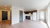 This single-floor unit presents a clean, contemporary design with light hardwood-style flooring flowing consistently throughout the space. The open-concept layout connects the kitchen and living area seamlessly, creating a functional and airy environment.

The kitchen is equipped with dark espresso cabinetry extending to the ceiling, offering generous storage. White appliances — including a refrigerator, range, microwave, and dishwasher — provide a crisp contrast against the dark cabinetry. Light countertops and a stainless steel sink complete the practical workspace. A louvered interior door adds a subtle design detail near the kitchen transition.

The living area is notably bright, benefiting from large floor-to-ceiling windows that frame urban rooftop views and flood the space with natural light. Recessed lighting supplements the natural illumination throughout.

The bathroom features warm-toned hardwood-style flooring, a full soaking tub with shower and curtain rod, a toilet, and a wide vanity with dark cabinetry matching the kitchen, topped with a light countertop and single sink. A large mirror spans the vanity wall, enhancing the sense of depth. The overall palette is neutral and modern, with white walls and understated finishes throughout.