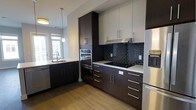 This single-floor space showcases a thoughtfully designed modern kitchen that balances aesthetics with practicality. The kitchen features a striking two-tone cabinetry scheme, pairing deep espresso-toned lower cabinets and tall pantry units with crisp white upper cabinets, creating a bold yet sophisticated contrast. Brushed silver bar-pull hardware adds a refined, contemporary touch throughout.

The cooking area is anchored by a built-in double wall oven and a flush cooktop, complemented by an integrated range hood. A dark subway tile backsplash serves as a standout design element, adding texture and depth to the space. Light quartz countertops provide a clean, bright work surface that contrasts beautifully against the darker cabinetry.

On the opposite wall, a stainless steel sink is positioned beneath a window that invites natural light into the kitchen, enhancing the overall brightness of the space. A stainless steel dishwasher and refrigerator complete the suite of appliances, ensuring a cohesive, professional-grade kitchen environment. Warm hardwood flooring runs throughout, grounding the space and adding an inviting element to the otherwise sleek, modern design.