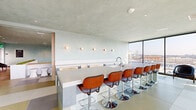 Located at 200 Mesnager St in Los Angeles, this sophisticated rooftop amenity space offers a seamless blend of indoor comfort and outdoor living. The interior is defined by soft sage-toned textured walls, large-format tile flooring in warm neutral tones, and recessed lighting that lends a calm, polished atmosphere throughout.

The first living area features a sleek, elongated bar counter with a built-in sink, lined with mid-century-inspired walnut and leather barstools on chrome pedestals. Circular wall sconces add a refined decorative touch, while floor-to-ceiling glass walls flood the space with natural light and frame expansive views of the Los Angeles skyline.

The second living area centers on a sunken lounge with wrap-around built-in bench seating, accented by a living moss-topped partition wall. Low-profile floor cushions and small round wood tables create an inviting, informal gathering space, complemented by additional lounge chairs positioned to take in the city views.

The outdoor terrace extends the space significantly, featuring a central fire pit surrounded by vibrant red chairs, pergola-shaded dining tables, and lush planters. Glass railings ensure unobstructed panoramic views of the downtown Los Angeles skyline and surrounding hillsides, making this rooftop an exceptional destination for both relaxation and entertainment.