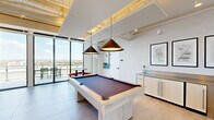 This impressive single-floor amenity space is thoughtfully designed to deliver a premium social and recreational experience. The open, airy layout is anchored by floor-to-ceiling glass walls and sliding doors that flood the interior with abundant natural light and frame sweeping panoramic views of the surrounding cityscape and beyond.

The game and recreation area serves as a central highlight, featuring a full-size billiards table beneath striking conical pendant lights, a foosball table, and a bank of classic arcade machines — all set against exposed ductwork ceilings with warm accent tones that lend an industrial-chic character. Adjacent lounge zones offer plush, curated seating arrangements, including bold statement chairs and curved sofas, arranged around flat-screen televisions and slatted wood accent walls.

A full-service bar and kitchen area showcases a dramatic waterfall-edge stone island, warm wood cabinetry with an arched architectural detail, and bar seating, making it ideal for entertaining. The space also connects seamlessly to a furnished outdoor terrace, equipped with high-top tables and seating, providing an elevated al fresco setting with unobstructed skyline views. Porcelain tile flooring runs throughout, tying together the various zones into a cohesive and sophisticated environment.