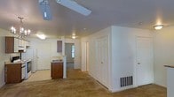 This single-floor residential space offers a practical, open layout that connects the main living area to the kitchen, creating a cohesive and functional floor plan. The living area features wall-to-wall carpeting in a neutral tone, providing a warm underfoot feel, and is equipped with a ceiling fan that aids in air circulation throughout the space. A floor-level HVAC vent is integrated into the baseboard, contributing to the home's climate control system. The kitchen is positioned toward the rear of the open area and features dark wood cabinetry, a sink, and a countertop workspace. A window above the kitchen sink allows natural light to filter into the space, brightening the interior. White six-panel interior doors are present throughout, lending a clean and classic aesthetic to the home. The walls are finished in a neutral white, complementing the overall understated palette. Multiple doorways branch off from the main living corridor, suggesting additional rooms such as a bathroom and at least one bedroom. The ceilings are of standard height, and flush-mount light fixtures provide consistent overhead lighting. Overall, the space presents a straightforward, move-in-ready layout suited for everyday residential living.