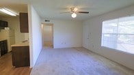 This single-floor space features a straightforward, functional layout with clearly defined areas. The main living area is finished with neutral-toned carpet, creating a soft underfoot feel, while the adjacent kitchen and connecting areas transition to hardwood-style flooring. A low partial wall divides the living space from the kitchen area, maintaining an open and connected feel without fully merging the two zones.

The kitchen area is equipped with wooden cabinetry in a warm brown finish, providing practical storage. A wall-mounted HVAC unit is visible, ensuring climate control throughout the space. The walls are painted in a clean, neutral white, contributing to a bright and airy atmosphere.

A hallway extends from the main living area, leading to additional rooms. Ceiling-mounted lighting fixtures are present, offering general illumination. The consistent use of neutral colors across walls and flooring gives the space a cohesive and understated aesthetic, making it adaptable for a variety of furnishing styles. Overall, the layout is practical and efficiently organized across its single floor.