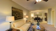 This thoughtfully designed single-floor residence offers a seamless blend of comfort and elegance. The open-concept layout connects the living and dining areas, creating a spacious and inviting atmosphere ideal for both relaxing and entertaining.

The living area is anchored by a plush cream leather sofa and coordinating armchair, arranged around a rustic wood coffee table that adds a natural, organic touch. A large landscape painting in rich earth tones serves as a striking focal point on the softly painted walls, while a ceiling fan overhead ensures year-round comfort. Warm ambient lighting from table lamps creates a cozy, welcoming glow throughout the space.

Flowing naturally from the living area, the dining space accommodates a full dining set with dark wood chairs and a statement chandelier that adds a refined, classic touch. A complementary piece of wall art echoes the nature-inspired aesthetic carried throughout the home.

The overall ambiance is warm and sophisticated, with a cohesive color palette of neutral tones accented by rich wood finishes and carefully chosen décor. The thoughtful arrangement of furniture and lighting throughout the space reflects an attention to detail that makes this residence both functional and visually appealing.