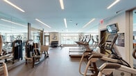 Located at 8815 Peregrine Heights Rd in Manassas, Virginia, this well-appointed fitness center offers a comprehensive workout environment across a single thoughtfully designed floor. The main exercise area is bright and spacious, illuminated by sleek linear ceiling lights that complement the contemporary aesthetic. Large windows line one wall, providing abundant natural light and direct views of the outdoor pool and lounge area, creating an energizing atmosphere for workouts.

The cardio zone is well-equipped with a row of treadmills and elliptical machines, while a dedicated section houses cable and multi-functional strength machines alongside free weights, including a full dumbbell rack. Striped flooring in this zone adds a modern, dynamic visual element. Mirrored walls expand the sense of space and support form monitoring during exercise.

A separate studio room features light wood flooring, warm orange accent walls, and wall sconces flanking a large built-in flat-screen television. This area is stocked with stability balls, medicine balls, and resistance equipment for functional and flexibility training.

An adjacent entry or transition area includes built-in shelving and storage cubbies with a glass partition wall, offering an organized space to store belongings before entering the workout floor. Together, these zones create a fully equipped and visually polished fitness facility.