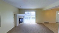 This single-floor residence offers a comfortable and functional layout designed for easy living. The living area serves as the heart of the home, highlighted by a classic white-mantled fireplace with a glass-front surround, adding both warmth and visual appeal to the room. A large sliding glass door floods the space with abundant natural light, offering a direct view of the lush greenery outside and providing seamless access to the outdoors. Vertical blinds on the sliding door allow for flexible light control throughout the day. The room is finished with plush wall-to-wall carpeting in a neutral tone, contributing to a soft and inviting ambiance. The walls are painted in a warm, light yellow hue that complements the natural light and enhances the open feel of the space. A built-in shelf or ledge on one wall provides a subtle architectural detail and practical display space. Baseboard heating vents are thoughtfully placed throughout, ensuring consistent comfort. Overall, the space presents a clean, well-maintained, and move-in-ready environment with a calm and livable character.