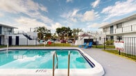 Located in Deerfield Beach, Florida, this outdoor pool deck serves as the central amenity of a two-story residential complex. The centerpiece of the space is a generously sized rectangular swimming pool with clear, blue-tinted water, metal handrails for safe entry, and depth markings along its perimeter. The pool is surrounded by a wide concrete deck that provides ample space for lounging, complemented by poolside seating such as a blue lounge chair.

The complex features a classic Florida architectural style, with white and light gray exterior walls accented by navy blue doors, railings, and window trim. Units on both the ground and upper floors face inward toward the courtyard, with the upper level accessed via exterior staircases. Open-air balconies line the upper floor, offering residents direct views of the pool area below.

The courtyard is enclosed by black iron fencing, which defines the pool area while maintaining visibility to the surrounding landscape, including mature trees and greenery beyond the perimeter. A life ring is mounted near the pool, and mailbox units are positioned along one edge of the fence line. The open-sky setting allows for abundant natural light throughout the day, creating a bright and inviting outdoor environment well-suited to Florida's sunny climate.