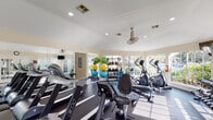 Located at 1202 Seagler Rd in Houston, TX, this well-appointed fitness center is designed to support a comprehensive workout experience within a bright, welcoming environment. The single-floor facility features vaulted ceilings that lend an open, airy feel to the space, complemented by large windows along multiple walls that allow abundant natural light to flood in while offering views of the surrounding greenery outdoors.

The gym is equipped with a diverse array of cardio machines, including treadmills, elliptical trainers, and a stationary recumbent bike, all arranged to maximize floor space and usability. A multi-station cable and weight machine anchors the strength training area, accompanied by a dedicated free weights section featuring dumbbells and a well-stocked rack of medicine balls and stability balls in assorted sizes, catering to a variety of fitness routines.

Dark rubber flooring throughout the main workout area provides both durability and comfort underfoot, while large wall-mounted mirrors enhance the sense of space and allow users to monitor their form. A ceiling fan ensures comfortable airflow during intense sessions. The overall ambiance is clean, functional, and motivating, making this fitness center a practical and well-equipped amenity for active individuals.