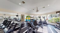Located at 1202 Seagler Rd in Houston, TX, this well-appointed fitness center is designed to support a comprehensive workout experience within a bright, welcoming environment. The single-floor facility features vaulted ceilings that lend an open, airy feel to the space, complemented by large windows along multiple walls that allow abundant natural light to flood in while offering views of the surrounding greenery outdoors.

The gym is equipped with a diverse array of cardio machines, including treadmills, elliptical trainers, and a stationary recumbent bike, all arranged to maximize floor space and usability. A multi-station cable and weight machine anchors the strength training area, accompanied by a dedicated free weights section featuring dumbbells and a well-stocked rack of medicine balls and stability balls in assorted sizes, catering to a variety of fitness routines.

Dark rubber flooring throughout the main workout area provides both durability and comfort underfoot, while large wall-mounted mirrors enhance the sense of space and allow users to monitor their form. A ceiling fan ensures comfortable airflow during intense sessions. The overall ambiance is clean, functional, and motivating, making this fitness center a practical and well-equipped amenity for active individuals.