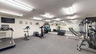 Located at 151 North St in Portland, Maine, this dedicated fitness center offers a well-equipped exercise space designed to support a comprehensive workout routine. The room features durable grey carpet flooring throughout and is illuminated by rows of bright fluorescent overhead lighting that keeps the space well-lit and energizing.

A full wall of mirrors along one side of the room amplifies the sense of space and allows users to monitor their form during exercise. The cardio equipment selection includes a treadmill, an elliptical trainer, a stationary upright bike, and a spinning bike, providing a variety of options for cardiovascular training. Strength training is well covered with a multi-station cable machine, an adjustable weight bench, and a full rack of dumbbells in graduated sizes. A medicine ball rack and a collection of fitness balls round out the functional training offerings.

A wall-mounted fan provides supplemental air circulation, and a flat-screen television is mounted on the wall for entertainment during workouts. A body weight scale is also available. Coastal-themed artwork decorates the white walls, adding a touch of local character reflective of Portland's maritime heritage. A water dispenser is accessible near the entry door, ensuring hydration is always within reach. The space is thoughtfully organized to maximize usability across its compact footprint.